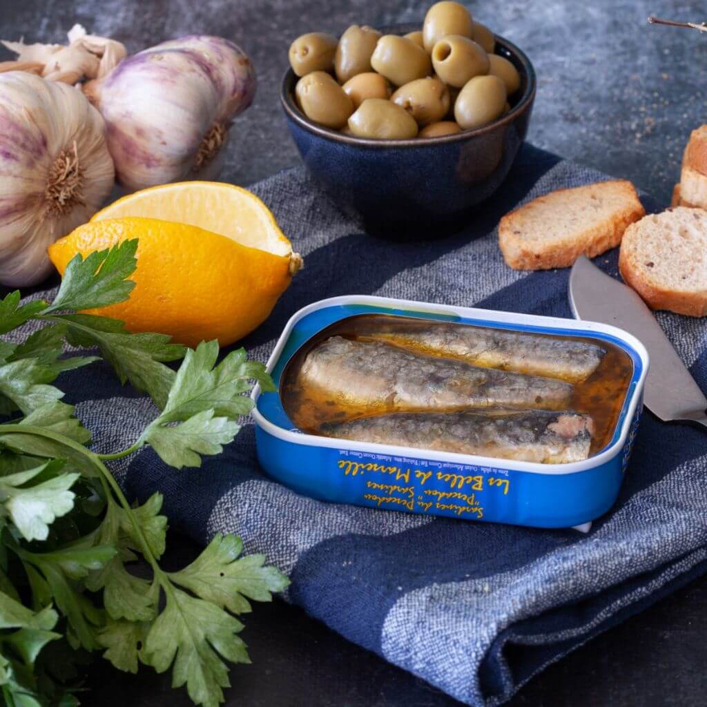 Eine offene Dose mit Ferrigno Sardinen Fischer Art mit Knoblauch, Zitrone & Petersilie. Angerichtet mit Weißbrot und grünen Oliven in Mannheim.