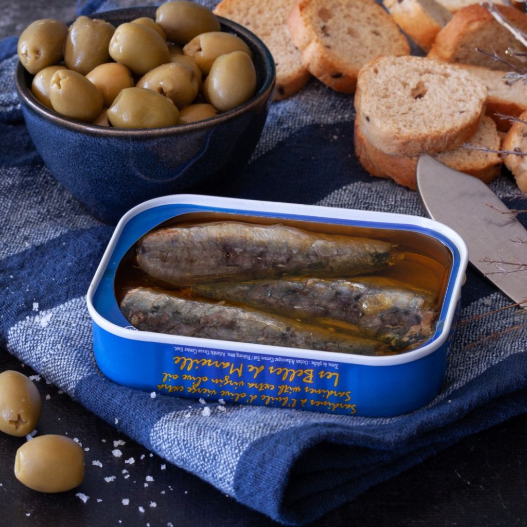 Eine offene Dose Ferrigno Sardinen in Olivenöl der Serie La Bonne de Marsaille n Mannheim mit Weißrot und einer Schüssel grüner Oliven.