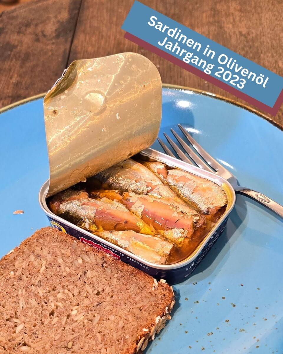 Geöffnete Jahrgangssardinen 2023 in Olivenöl mit feinen Sardinen. Besonders aromatisch und zart im Geschmack. Blauer Teller mit Sardinendose und Brot. Feinkost online Mannheim