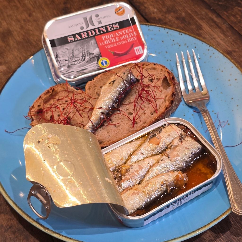 Ein blauer Teller mit einer offenen Dose Sardinen mit Chili und einer sScheibe rustikales Brot.