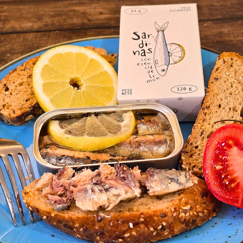 Geöffnete Dose der spanischen El Raño Sardinen mit Zitrone, angerichtet auf einer gerösteten Brotscheibe mit Tomate und Zitronenscheibe auf einem blauen Teller bei Feinkost Probieren & Genießen in Mannheim.