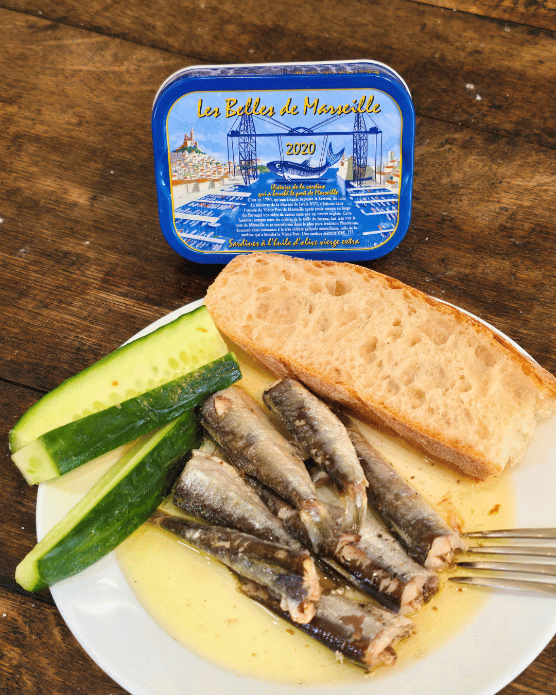 Ein Teller mit Sardinen in Olivenöl mit Baquette und grüner Gurke auf Holztisch. La Belle de Marsaille Jahrgangssardinen in Mannheim.
